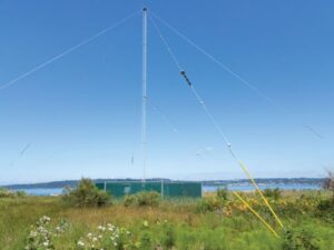 Tall communication tower with guy wires in a grassy field near water.